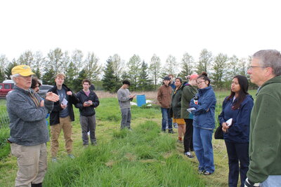 A group of people standing in a field listening to a man in a hat speak.