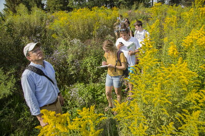 A man is looking at the sky in a field while students follow behind him writing in notebooks.