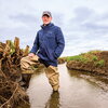 A man standing in a stream wearing a rain jacket, hat, and wader boots.
