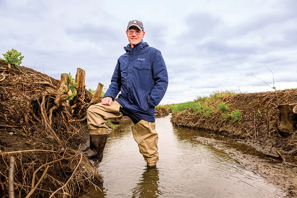 A man standing in a stream wearing a rain jacket, hat, and wader boots.