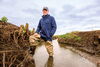 A man standing in a stream wearing a rain jacket, hat, and wader boots.